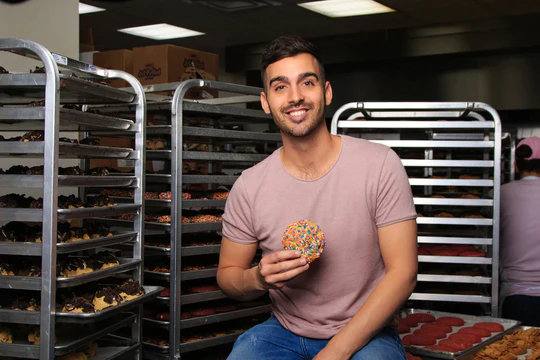 man sitting and holding a cookie with bakers rack behind